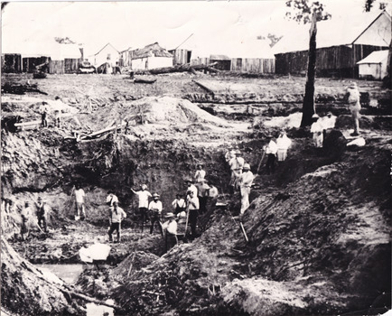 a black and white photograph depicting 22 men in a mining excavation area holding shovels etc. In the background, above the excavation pit, is a row of slab huts.