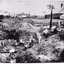 a black and white photograph depicting 22 men in a mining excavation area holding shovels etc. In the background, above the excavation pit, is a row of slab huts.