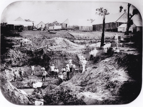 a black and white photograph depicting 22 men in a mining excavation area holding shovels etc. In the background, above the excavation pit, is a row of slab huts.