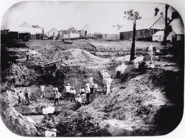 a black and white photograph depicting 22 men in a mining excavation area holding shovels etc. In the background, above the excavation pit, is a row of slab huts.