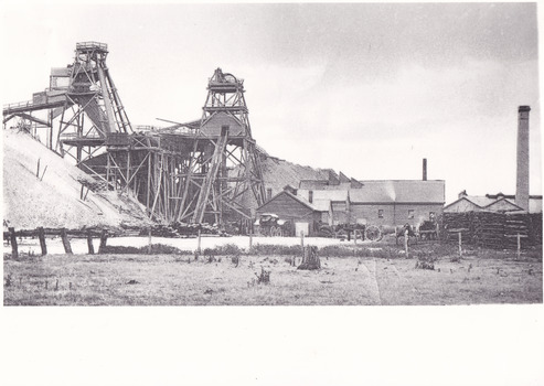 a black and white photograph of an early 1900s style mine, with buildings  and chimneys in the background.