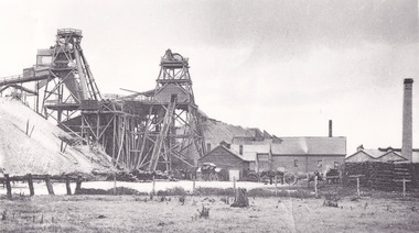 a black and white photograph of an early 1900s style mine, with buildings  and chimneys in the background.