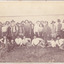 A image of a group of men and a few boys dressed for the time with hats on. Behind them is a tin fence. 