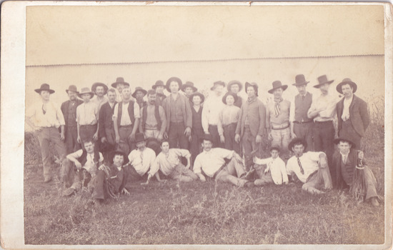 A image of a group of men and a few boys dressed for the time with hats on. Behind them is a tin fence. 
