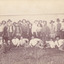 A image of a group of men and a few boys dressed for the time with hats on. Behind them is a tin fence. 