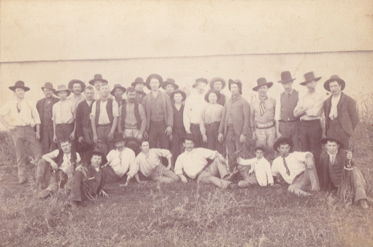 A image of a group of men and a few boys dressed for the time with hats on. Behind them is a tin fence. 