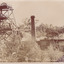A picture of a building in the middle ground on the right with a smoke stack to the left and a mining poppet head on the far left. A crowd of men are gathered in front of the building, standing near or sitting on a wood stack and looking at the camera. Another group of men are kneeling or standing underneath the poppet head structure. Numerous trees in the foreground and background. See transcript for text.