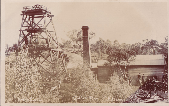 A picture of a building in the middle ground on the right with a smoke stack to the left and a mining poppet head on the far left. A crowd of men are gathered in front of the building, standing near or sitting on a wood stack and looking at the camera. Another group of men are kneeling or standing underneath the poppet head structure. Numerous trees in the foreground and background. See transcript for text.