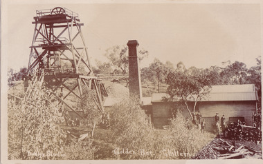 A picture of a building in the middle ground on the right with a smoke stack to the left and a mining poppet head on the far left. A crowd of men are gathered in front of the building, standing near or sitting on a wood stack and looking at the camera. Another group of men are kneeling or standing underneath the poppet head structure. Numerous trees in the foreground and background. See transcript for text.