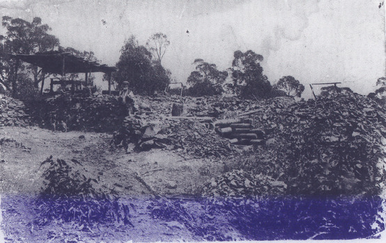 Scattered piles of smashed rock debris. A bearded man in a waistcoat stands near an open-sided timber structure. A man stands next to a dog below three seated men under a leaning four-posted structure. 