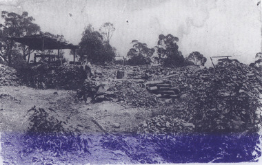 Scattered piles of smashed rock debris. A bearded man in a waistcoat stands near an open-sided timber structure. A man stands next to a dog below three seated men under a leaning four-posted structure. 