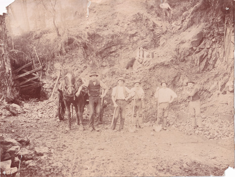 Seven miners and a horse and cart are depicted in front of mine a dug out hill. Five men stand alongside the horse facing the camera,  one man sits up the hill with a dog, and a final man stands higher. Six of the men lean against a pick or shovel, and six wear hats.  Broken rocks from the cliff are depicted strewn around the ground. 