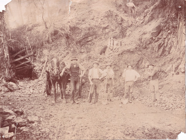 Seven miners and a horse and cart are depicted in front of mine a dug out hill. Five men stand alongside the horse facing the camera,  one man sits up the hill with a dog, and a final man stands higher. Six of the men lean against a pick or shovel, and six wear hats.  Broken rocks from the cliff are depicted strewn around the ground. 