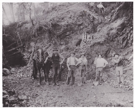 Seven miners and a horse and cart are depicted in front of mine a dug out hill. Five men stand alongside the horse facing the camera, one man sits up the hill with a dog, and a final man stands higher. Six of the men lean against a pick or shovel, and six wear hats. Broken rocks from the cliff are depicted strewn around the ground.