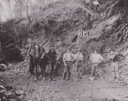 Seven miners and a horse and cart are depicted in front of mine a dug out hill. Five men stand alongside the horse facing the camera, one man sits up the hill with a dog, and a final man stands higher. Six of the men lean against a pick or shovel, and six wear hats. Broken rocks from the cliff are depicted strewn around the ground.