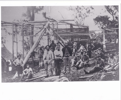 A black and white rectangular photograph depicting 21 separate men, either standing or sitting around a wooden structure.
