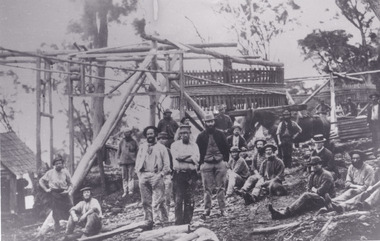 A black and white rectangular photograph depicting 21 separate men, either standing or sitting around a wooden structure.