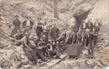 A sepia toned postcard depicting numerous men posing outside of a mineshaft. 