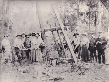 A black and white rectangular photograph depicting men and women standing around mining equipment. 