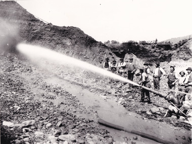 A black and white photograph featuring a group of ten miners standing at a cliff face using a high pressure hose.