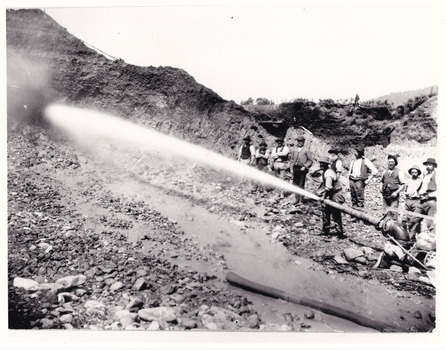 A black and white photograph featuring a group of ten miners standing at a cliff face using a high pressure hose.