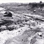 Black and white photograph showing a large excavation of a mining site. There are several figures in the foreground, and a pile of timber perhaps used for the mining infrastructure. There is a township sitting up on a hill in the distance in the background of the photograph and buildings on either side of the mining tailrace. 