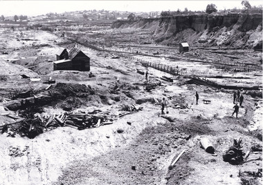 Black and white photograph showing a large excavation of a mining site. There are several figures in the foreground, and a pile of timber perhaps used for the mining infrastructure. There is a township sitting up on a hill in the distance in the background of the photograph and buildings on either side of the mining tailrace. 