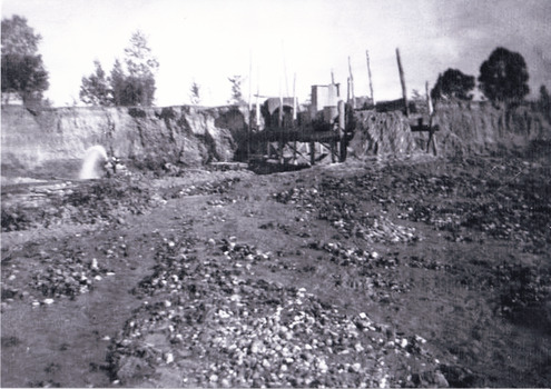 Scattered rocks and sluicing water jet flowing along a bank of earth eroded by sluicing machinery. A platform sits on timber posts in the rear with sluicing construction equipment. 