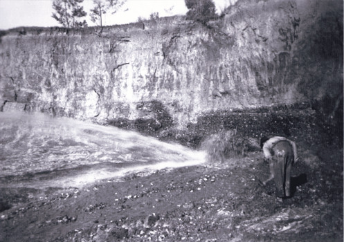 A black and white photograph depicting a man bending over and looking at the ground in a large mining cavity,. 