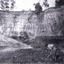 Man in black hat, white shirt and dark trousers bent over using a mining pick digging in the dirt in the foreground. Large rock wall in the background.