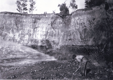 Man in black hat, white shirt and dark trousers bent over using a mining pick digging in the dirt in the foreground. Large rock wall in the background.