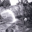 Hose spraying water in left foreground, man wearing dark trousers, a white shirt and a dark hat bent over using a pick digging up the dirt. A second man seen in the right background also digging dirt in the mine. Rock wall face of mine in the background with trees on top..