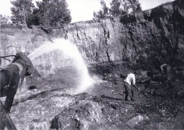 Hose spraying water in left foreground, man wearing dark trousers, a white shirt and a dark hat bent over using a pick digging up the dirt. A second man seen in the right background also digging dirt in the mine. Rock wall face of mine in the background with trees on top..