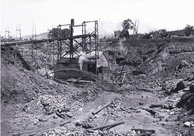 Open mine with bamboo scaffolding leading from left of photo to the centre. A small rectangular boiler/steamer house right behind scaffolding in centre of photo. Men with two horse and carts at top of mine at the right side of the background. 