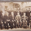 This photograph is framed in a cardboard mount and depicts a group of sixteen men in early 20th century attire standing and sitting in front of a stone building in the background. See the transcript of the reverse side of the image for names.