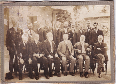 This photograph is framed in a cardboard mount and depicts a group of sixteen men in early 20th century attire standing and sitting in front of a stone building in the background. See the transcript of the reverse side of the image for names.
