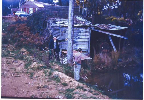 Two unknown men standing in front of a damn and next to an old wooden building that is becoming overrun by foliage. There is farm and perhaps mining equipment of some sort in an annex next to the building. Perhaps an old seam engine that has been converted to a water pump.