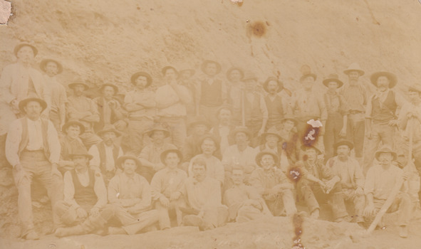 A faded sepia photograph depicting 34 men in work clothing in front of a bank of earth