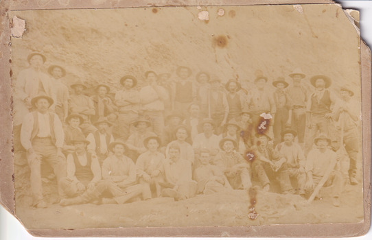 A faded sepia photograph depicting 34 men in work clothing in front of a bank of earth