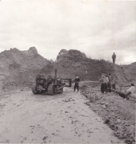 A black and white photograph. In the centre is a tractor. A person stands next to the tractor, showing it to a few onlookers gathered to the right.