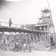 Black and white photograph with two men on wooden bridge structure and four men on ground below. Mining set up. 
