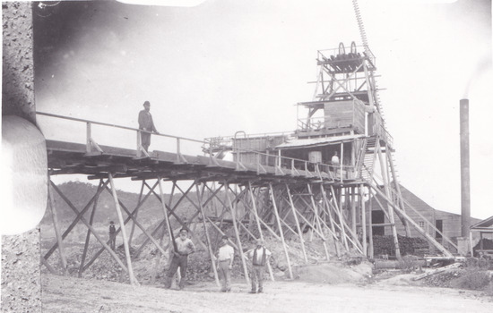 Black and white photograph with two men on wooden bridge structure and four men on ground below. Mining set up. 