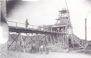 Black and white photograph with two men on wooden bridge structure and four men on ground below. Mining set up. 