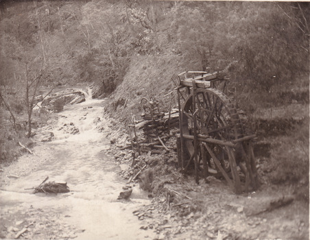 River flowing through scrub towards a metal wheel; part of a mine set up. (cropped)