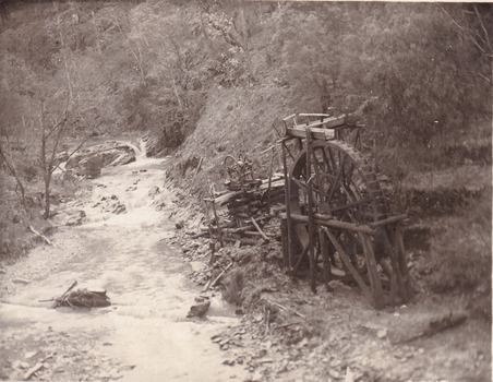 River flowing through scrub towards a metal wheel; part of a mine set up. (cropped)