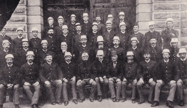 Black and white photo of 40 men in guard uniforms posing for a group photo in front of a building.