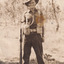 A sepia photo featuring an Australian World War I soldier dressed in army clothing, standing in the centre of the frame, holding a gun with a background of gum trees.