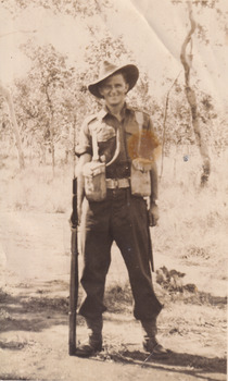 A sepia photo featuring an Australian World War I soldier dressed in army clothing, standing in the centre of the frame, holding a gun with a background of gum trees.
