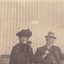 A sepia photo featuring a woman and a man, both older, seated together on a bench. The man wears a hat, full suit and glasses and the woman wears a hat and face veil and appears to be in widow’s mourning attire. 
