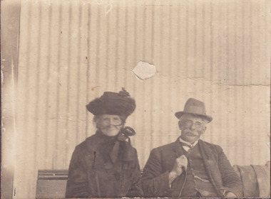 A sepia photo featuring a woman and a man, both older, seated together on a bench. The man wears a hat, full suit and glasses and the woman wears a hat and face veil and appears to be in widow’s mourning attire. 
